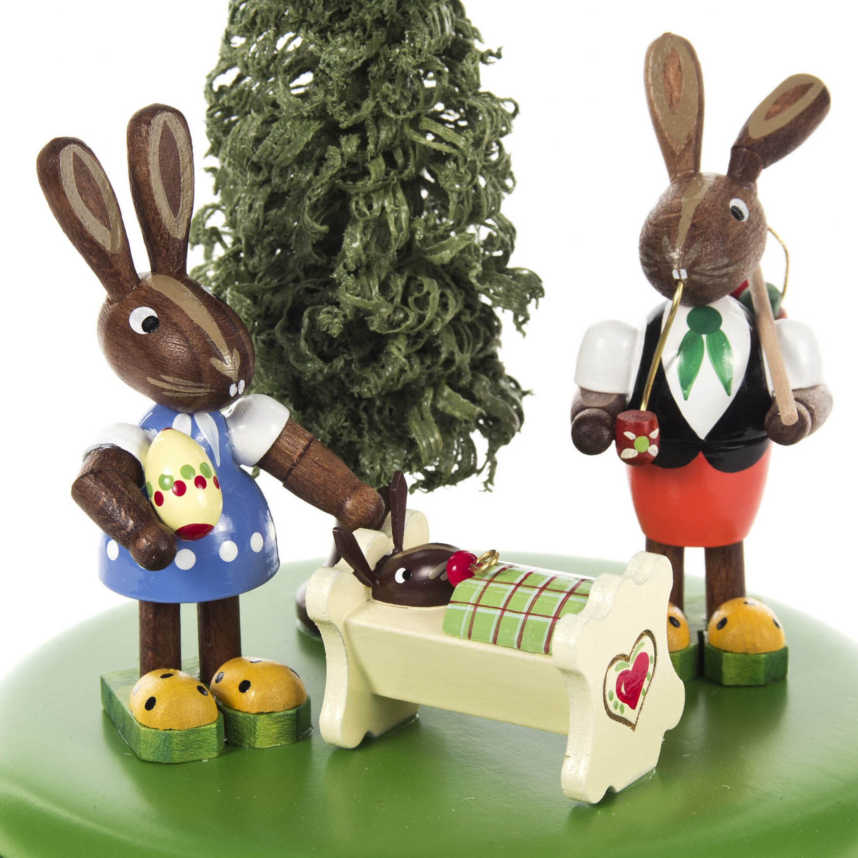Eine Spieldose mit zwei Holzosterhasen in traditioneller Kleidung; ein Hase steht mit einem Löffel in der Hand, während der andere einen in einer Wiege liegenden Hasen streichelt.