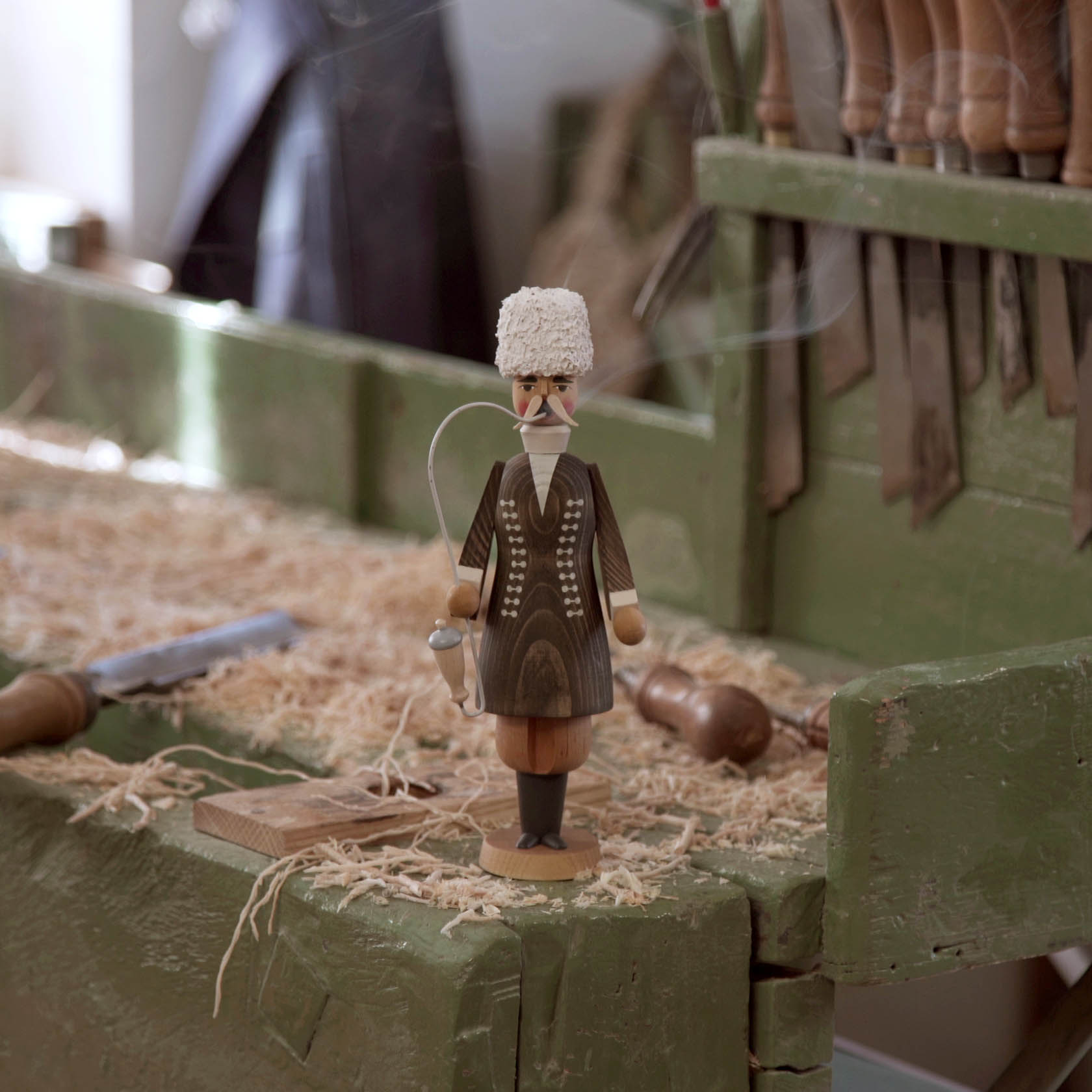 Ein traditioneller Räuchermann in der Kleidung eines Tscherkessians steht auf einer grünen Werkbank. Der handgeschnitzte Holzfigur trägt eine weiße Fellmütze und braune Kleidung.