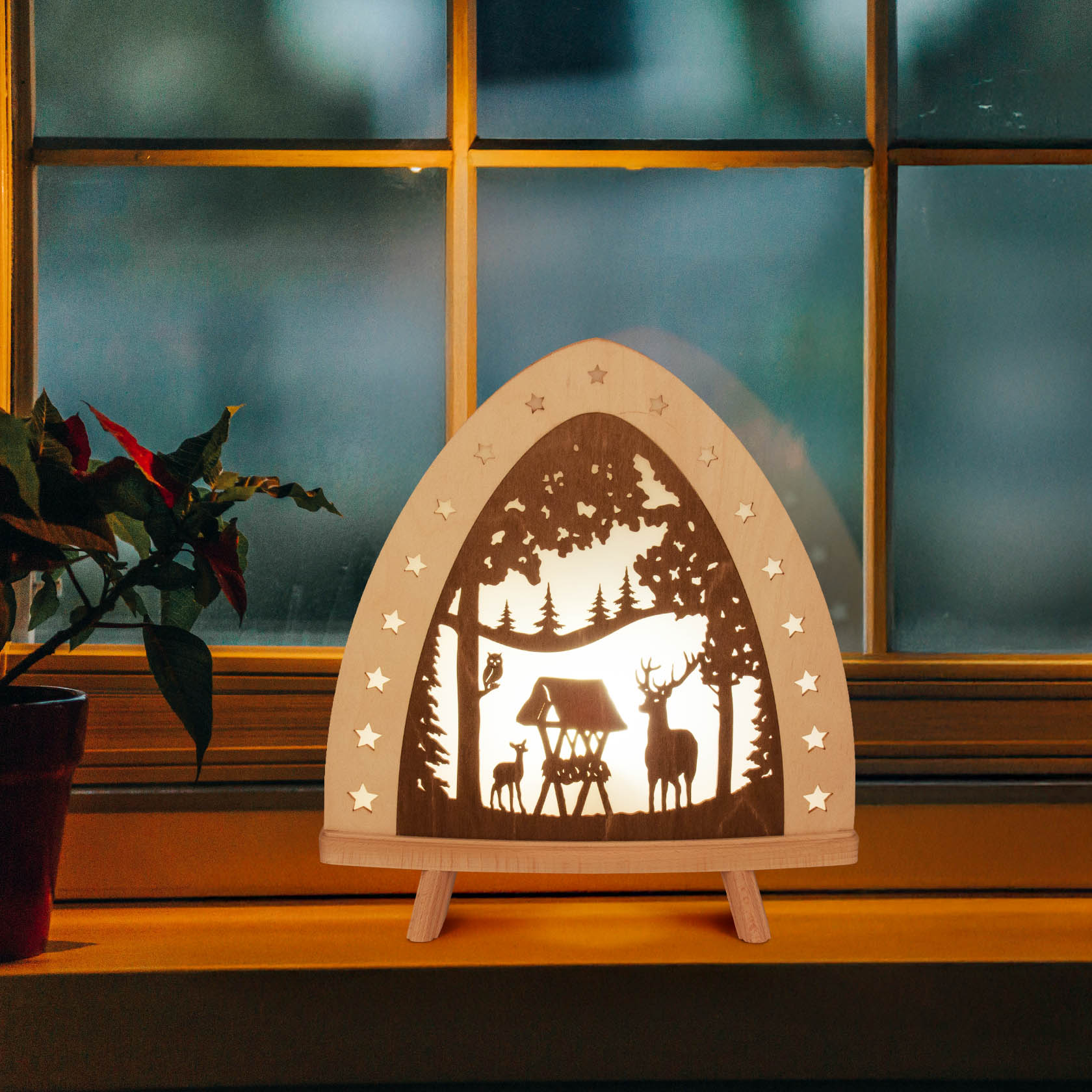 LED-Standleuchte mit Hirschmotiv zeigt Hirschfamilie und Bäume, umrandet von Sternen, auf einem Fensterbrett vor einem Fenster.