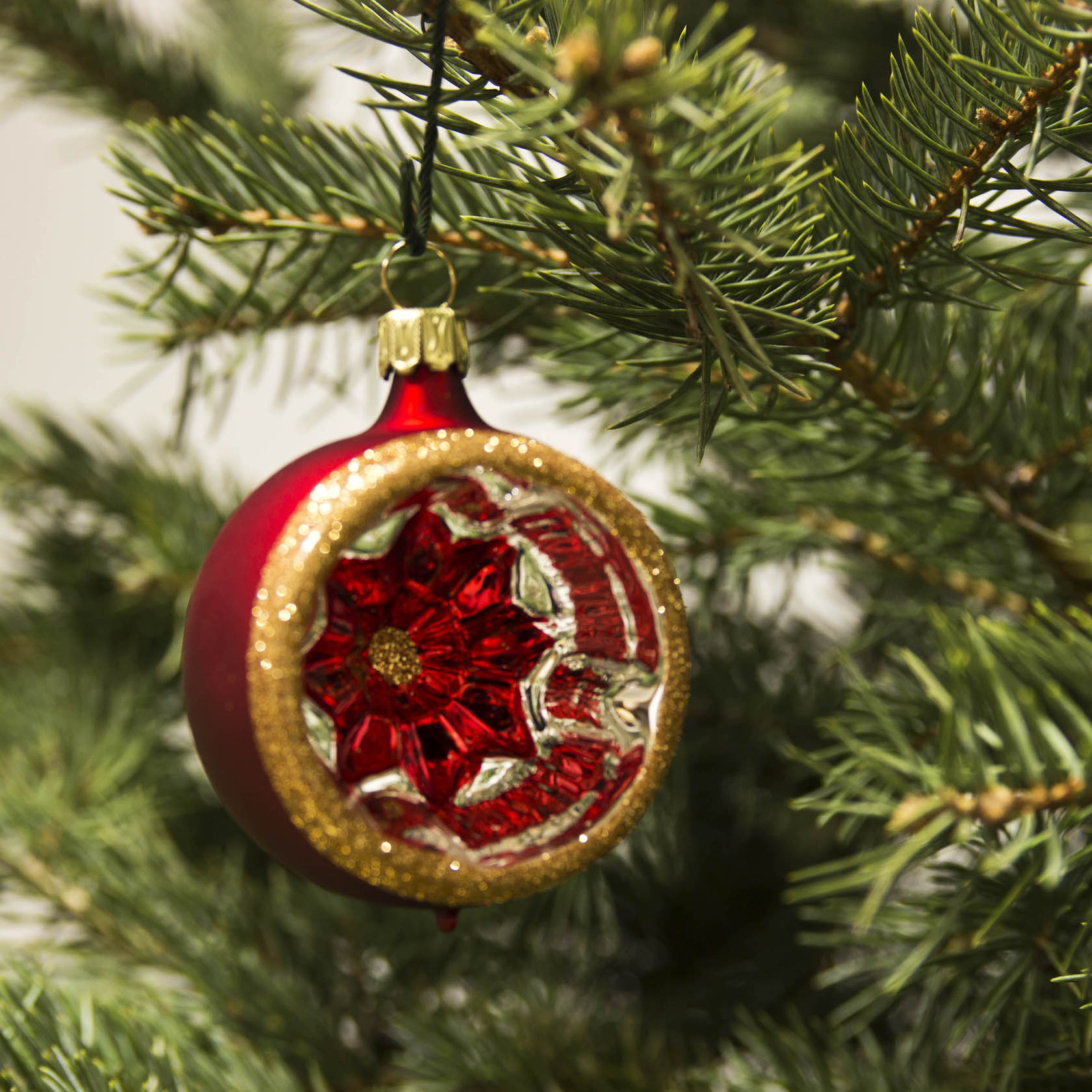 Eine rote, matte Christbaumkugel mit goldenen Glimmerverzierungen und einem reflektierenden Innenmuster hängt an einem Tannenzweig.