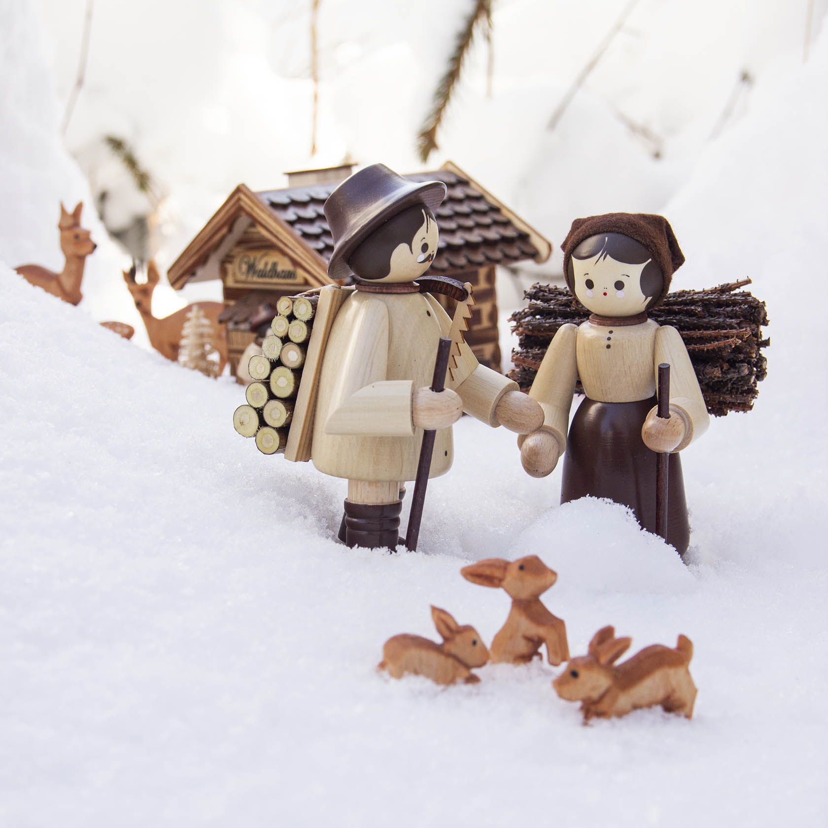 Zwei Holzfiguren im Schnee: Ein Mann mit Rückentrage voller Holzscheite und eine Frau mit Holzstapel auf dem Rücken. Drei kleine Häschen im Vordergrund.