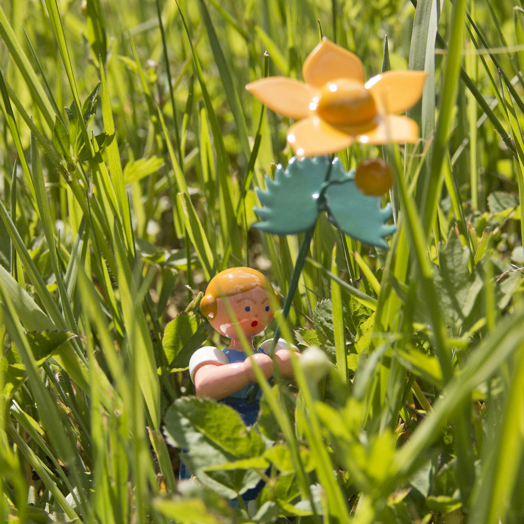 Holzfigur eines Mädchens in einem blauen Kleid mit einer gelben Sumpfdotterblume inmitten von grünem Gras.