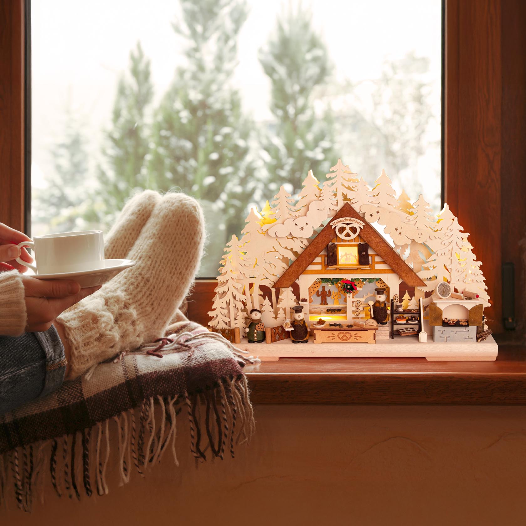 A small wooden Christmas arch with indirect electric lighting, depicting a cozy bakery scene, surrounded by snow-tipped trees and Christmas decorations.