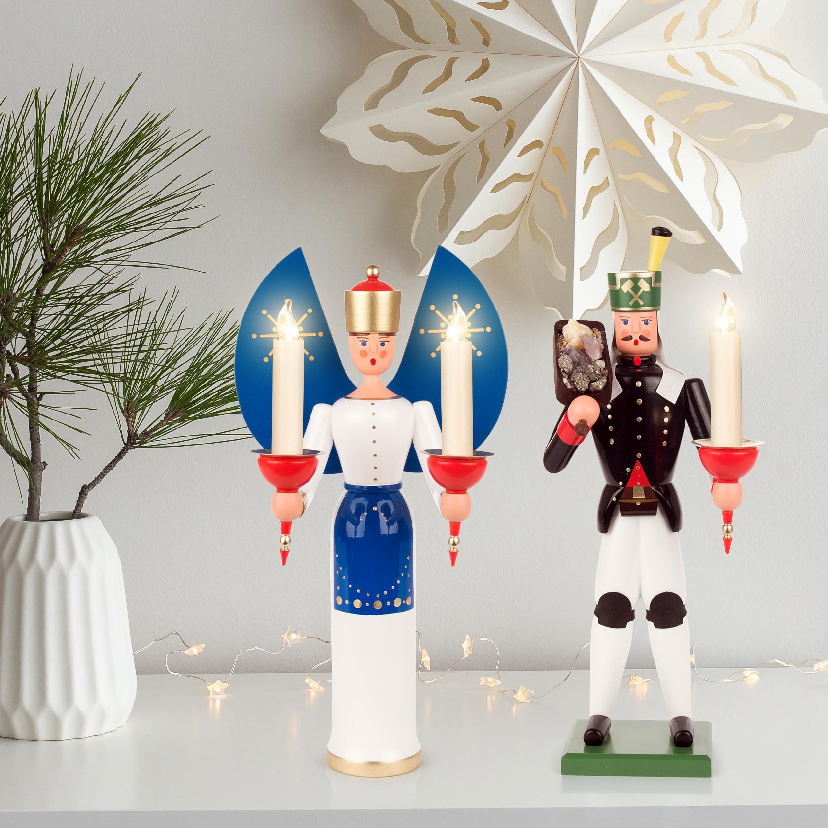 A wooden miner figure and an angel figure, both holding candle holders. The miner holds a mineral, while the angel holds two candles. Both stand on a shelf.