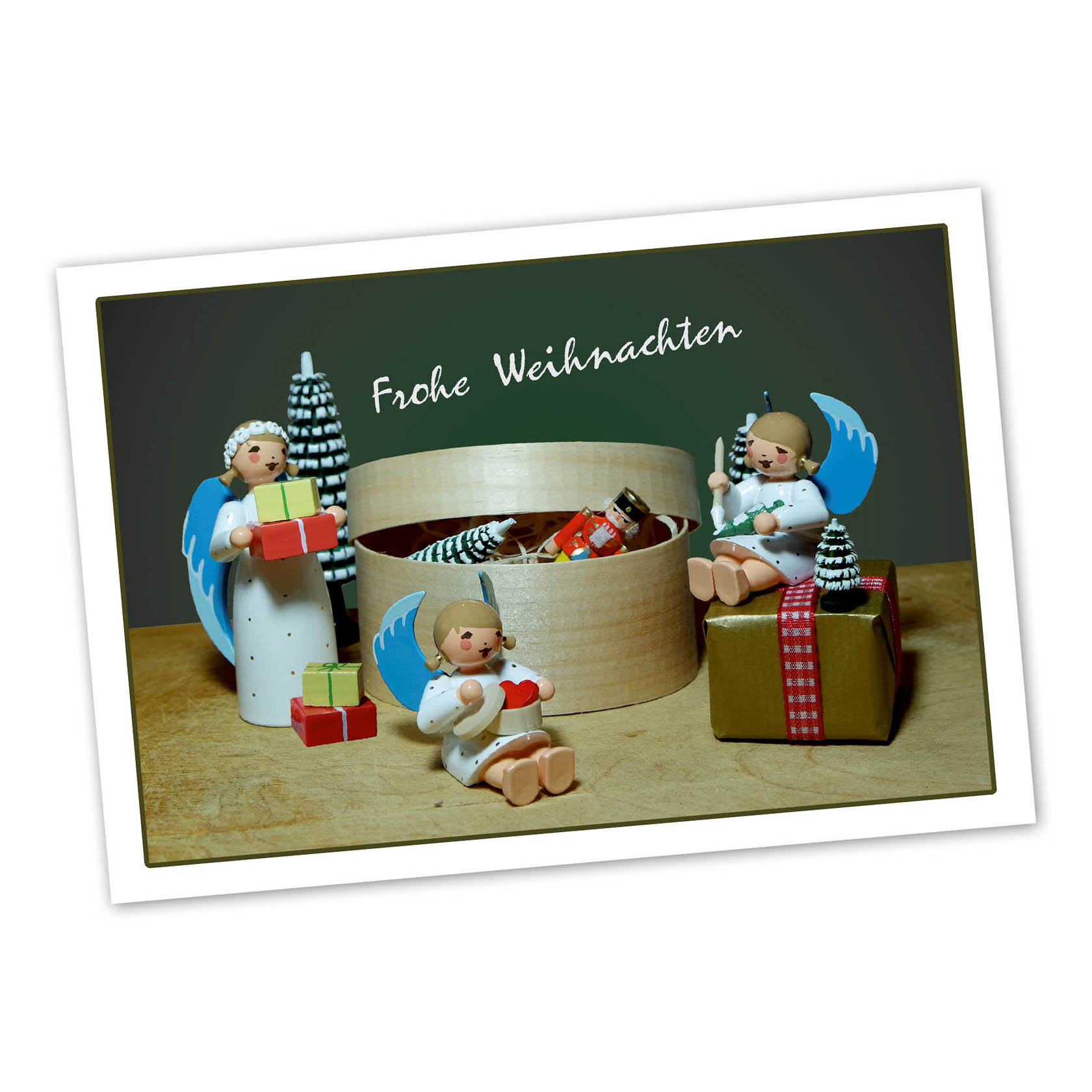 Three wooden angels with blue wings, holding gifts, surrounding a wooden box filled with small Christmas trees and candies. 'Frohe Weihnachten' text above. im Dregeno Online Shop günstig kaufen