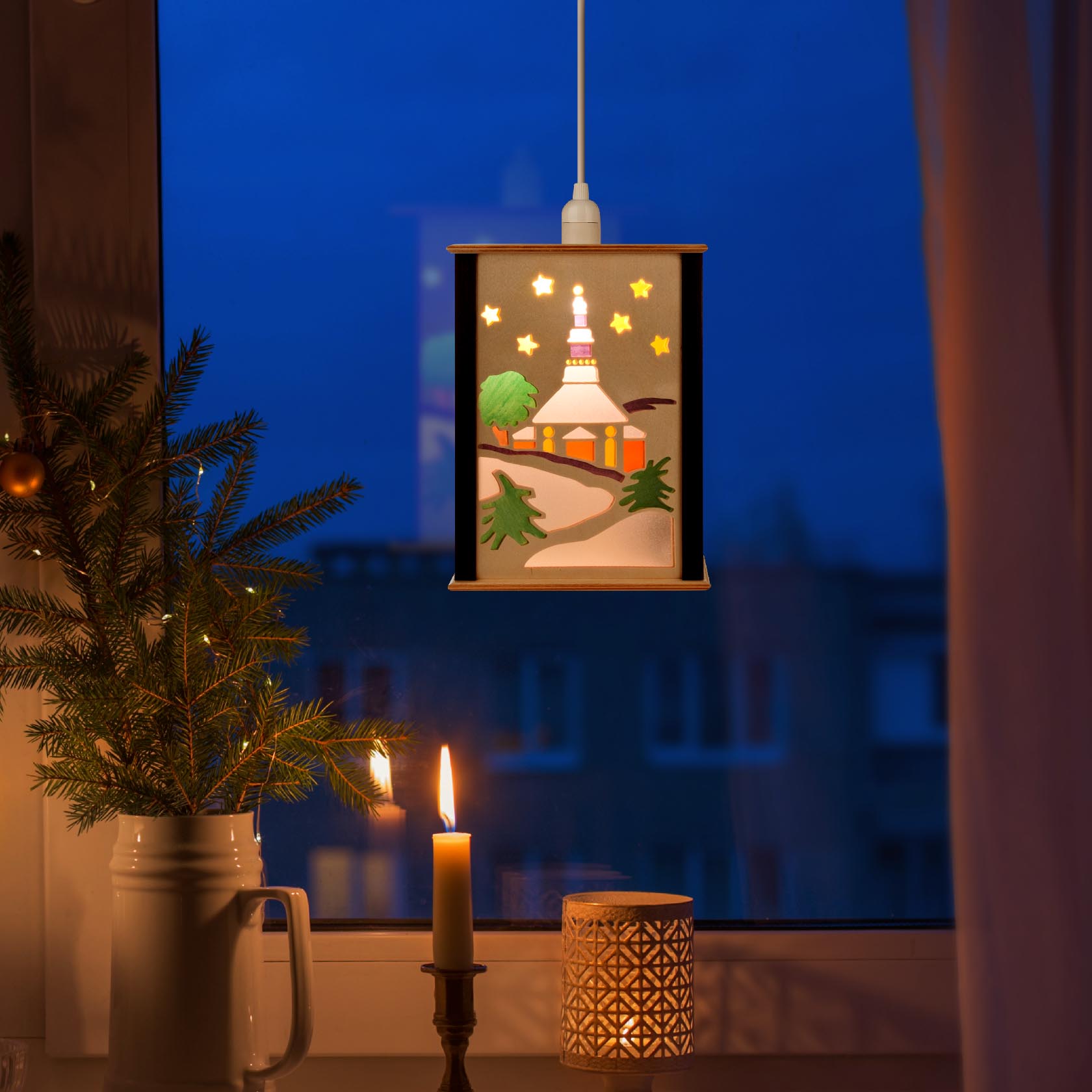 Eine beleuchtete Fensterlaterne zeigt eine Kirche mit Sternen, einem Tannenbaum und einem gebogenen Weg, aufgehängt vor einem Fenster bei Dunkelheit.