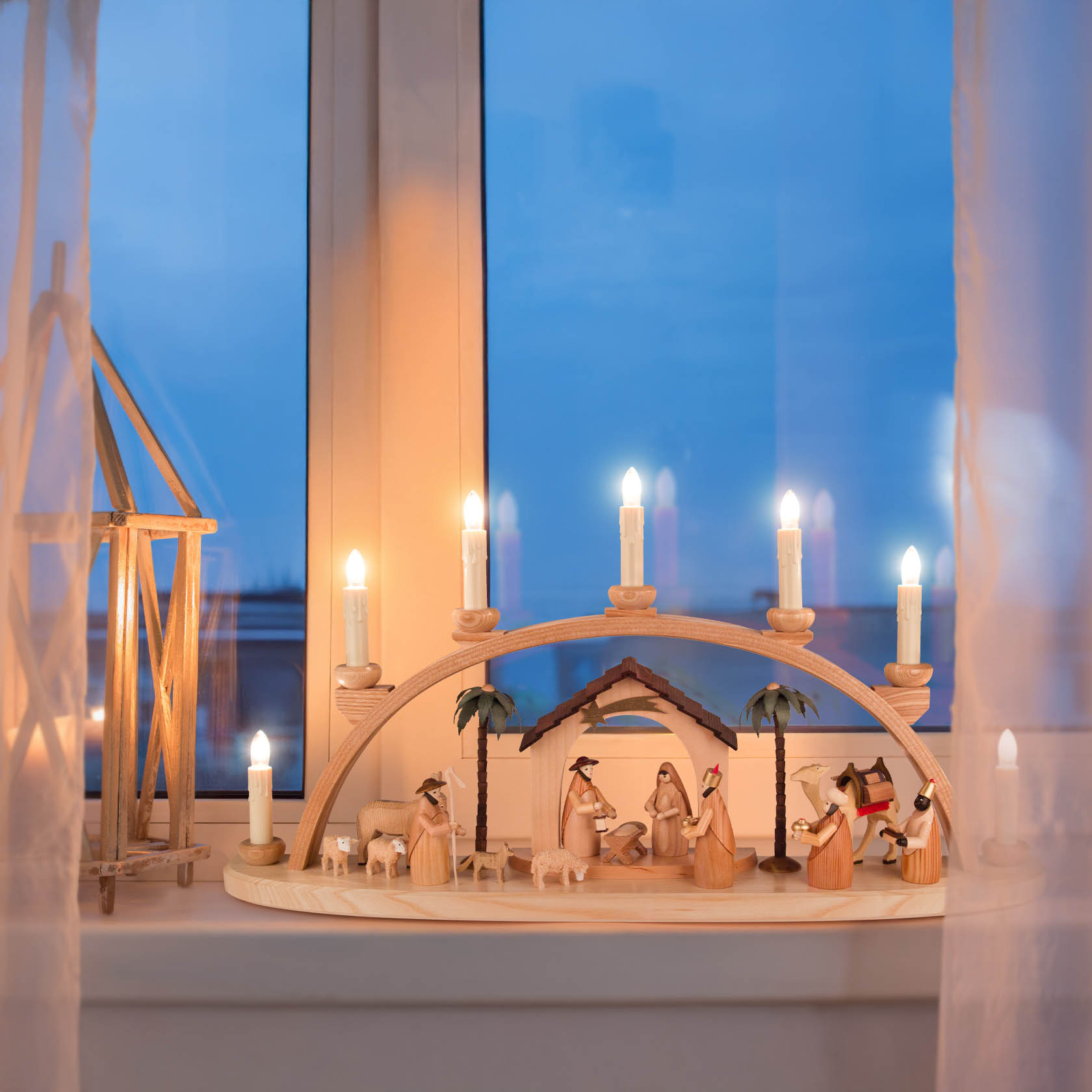 Ein natürlicher Schwibbogen mit der Szene der Geburt Christi, elektrisch beleuchtet. Figuren aus Holz, in einem Fenster aufgestellt.
