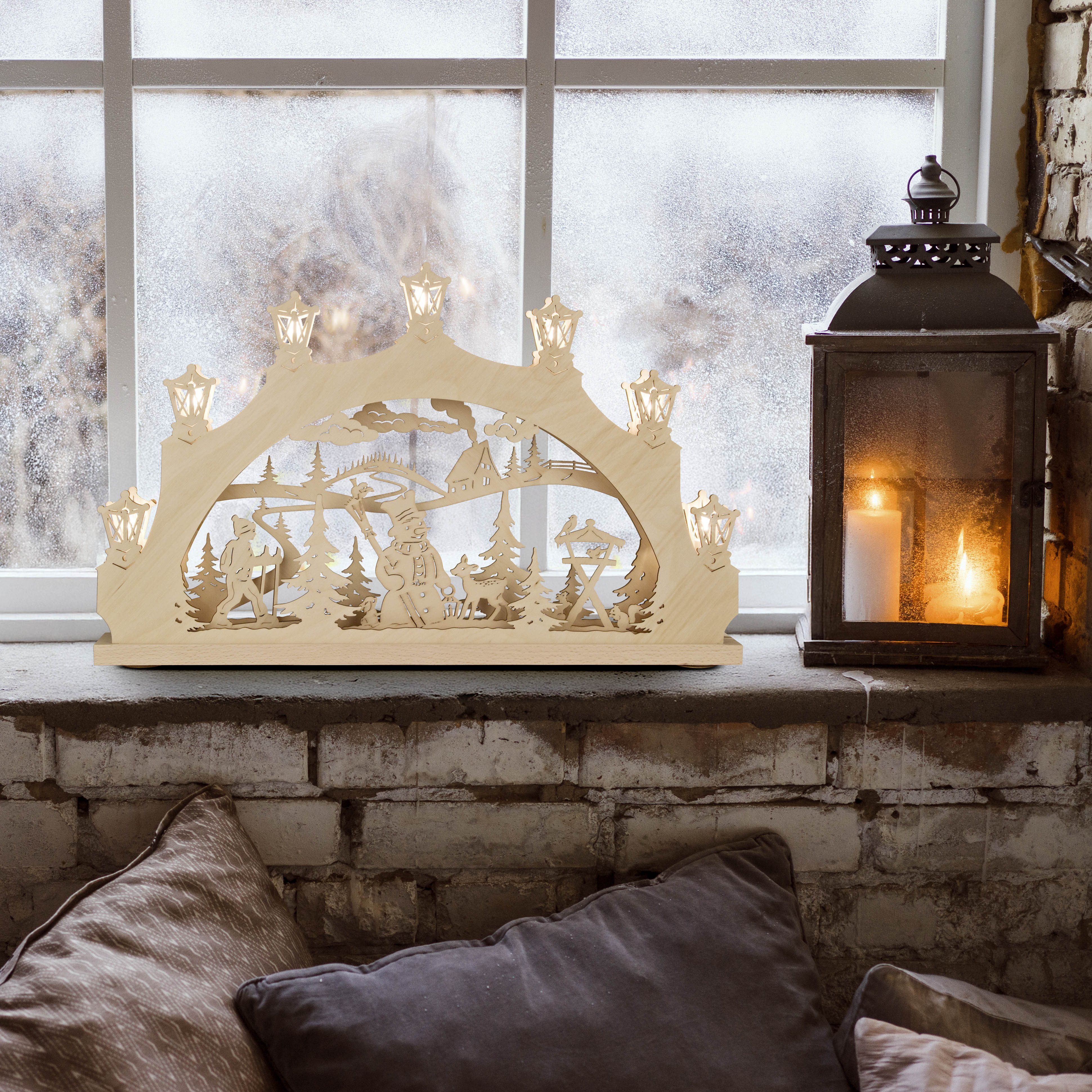 A wooden arch candle holder features a snowman in the forest scene with LED lights, placed on a windowsill. A lantern with candles is next to it.